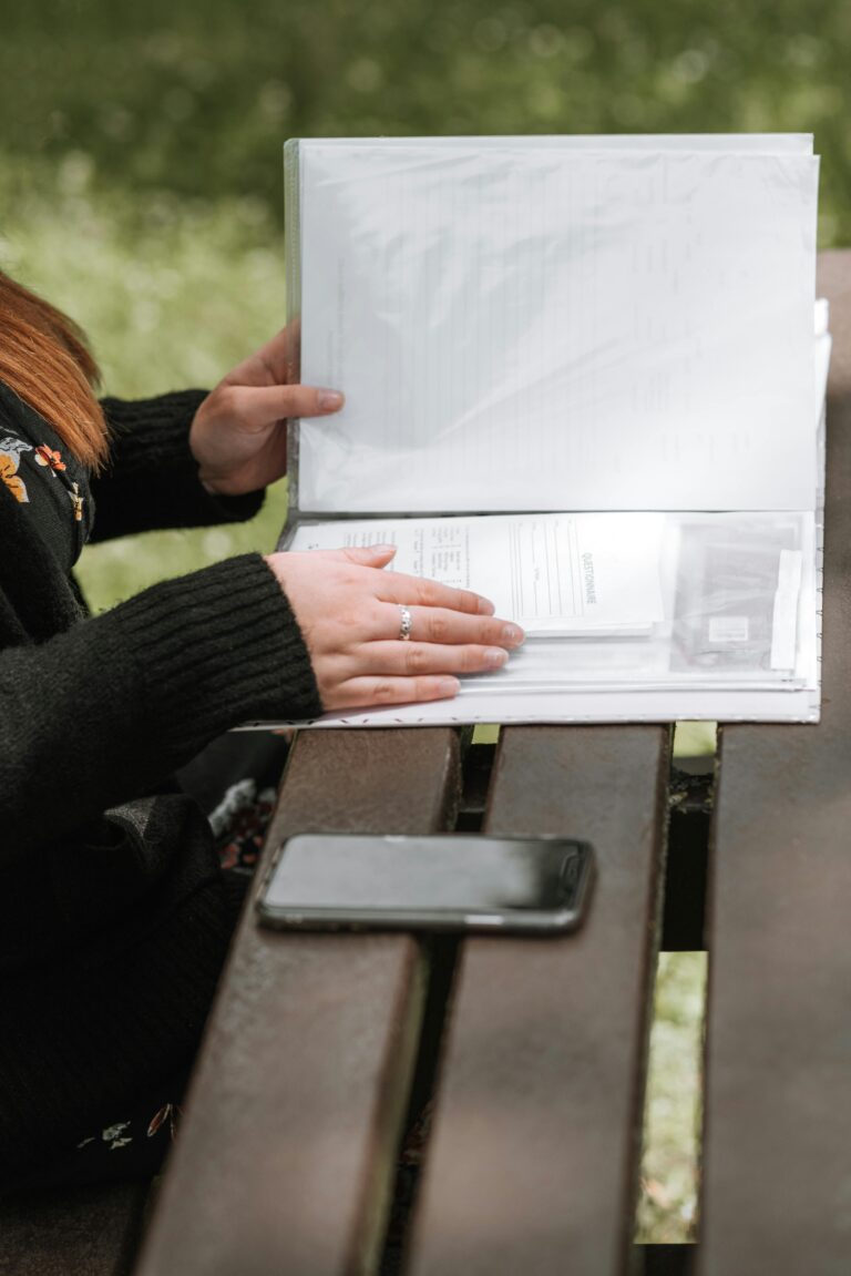 Side view crop anonymous female sitting at wooden table in green garden and reading papers in folder